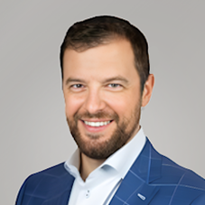 Dan Phillips A smiling man with short dark hair and a beard, wearing a blue checkered suit jacket over a white shirt, posed in front of a light gray background.