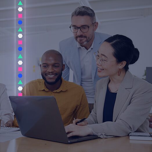 Three people collaborate around a laptop at a desk in a brightly lit office, with colorful geometric icons displayed vertically on the left side of the image.