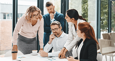 Five professionals gather around a table in a modern office, discussing documents and charts in a collaborative meeting.