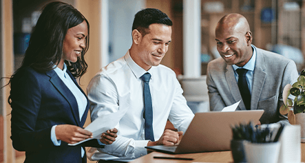Three business professionals in suits are reviewing documents and collaborating on a laptop with GridVizion in an office setting.