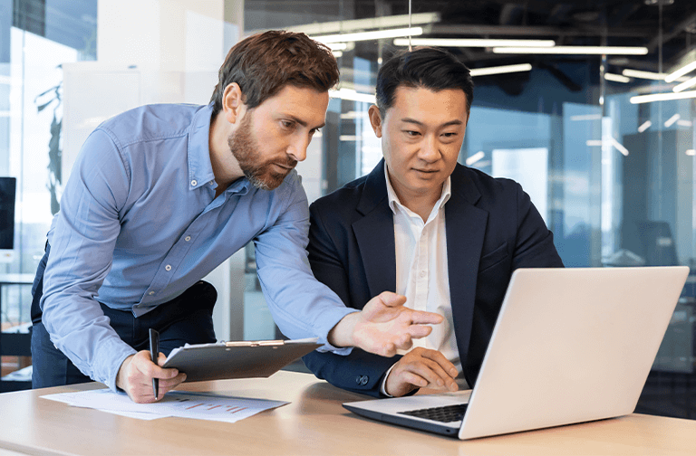 Two men in business attire are working together at a desk, one pointing at a laptop displaying GridVizion while the other holds a clipboard and pen.